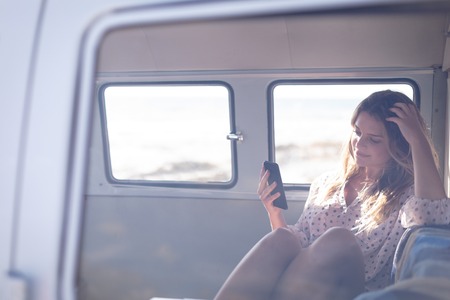 Front view of a beautiful thoughtful Caucasian woman using mobile phone in camper van at beach on a sunny dayの写真素材