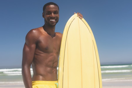 Portrait of handsome African American male surfer with a surfboard standing on a beach on a sunny...