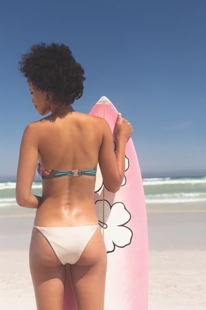 Rear view of fit young Mixed-race female surfer with a surfboard standing on a beach on a sunny day. Female looking to the left.の写真素材