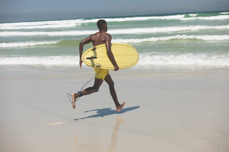 Rear view of handsome fit African American male surfer with a surfboard running towards the sea water on the beach on a sunny dayの写真素材