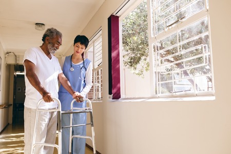Front view of Asian female nurse helping senior Caucasian male patient to walk with walker in corridor at retirement homeの写真素材
