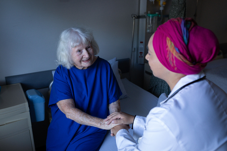 Front view of a mature mixed-race female doctor talking with a senior patient while holding her hands in a hospital roomの写真素材