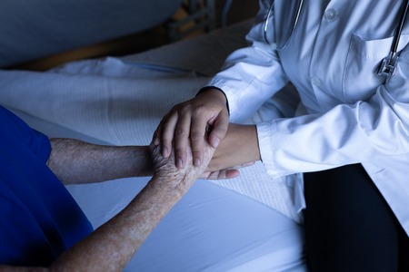 Mid section of a senior Caucasian female patient holding hands of a mixed race doctor in hospital roomの写真素材