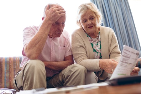 Front view of worried senior Caucasian couple discussing over medical bill while sitting on sofa at retirement home. Senior male has hand on his head.の写真素材