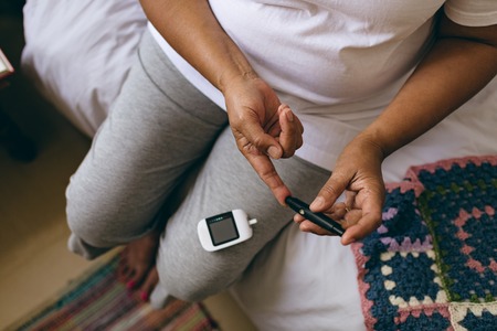 Low section of mixed race female senior patient checking blood sugar level with glucometer at retirement homeの写真素材