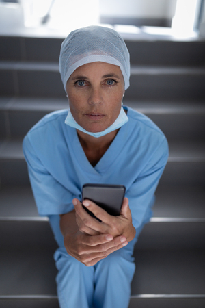 Portrait of a Caucasian female surgeon looking at the camera while sitting on stair case and holding mobile phoneの写真素材