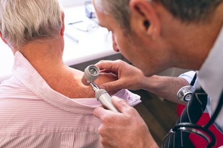 Front view of matured male Caucasian dermatologist examining senior patient through dermatoscopy at clinicの写真素材