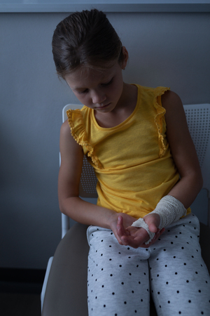 Front view of sad Caucasian girl sitting with fractured hand in clinic at hospitalの写真素材