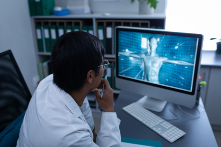Rear view of thoughtful confident Asian male doctor looking on computer in office at hospitalの写真素材