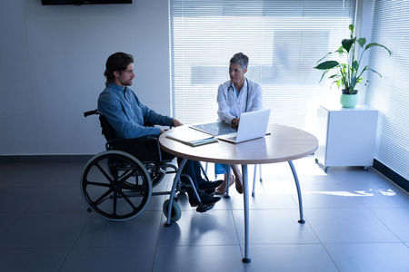 Front view of mature Caucasian female doctor and disabled Caucasian patient in wheelchair discussing over laptop in clinic at hospitalの写真素材