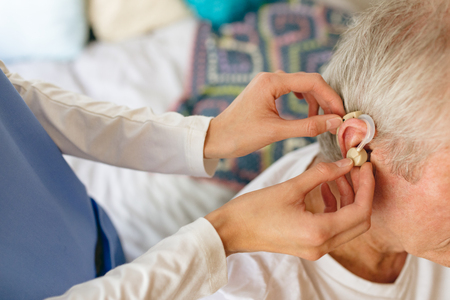 How angle view of female nurse applying hearing aid to senior Caucasian male patient ear at retirement homeの写真素材