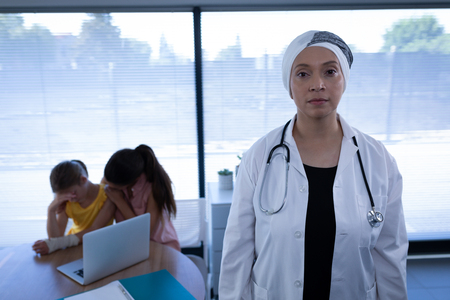 Front view of matured mixed race female doctor standing in clinic while Caucasian mother and daughter sitting upsetの写真素材