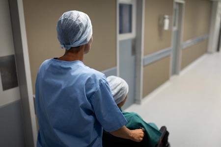 Rear view of a Caucasian female surgeon pushing her patient wheelchair through the hospital corridor
