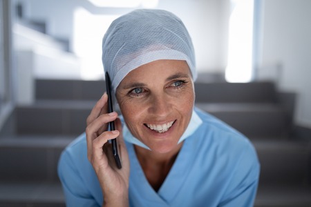 Front view of a Caucasian female surgeon smiling and talking on mobile phone while sitting on hospital stair caseの写真素材