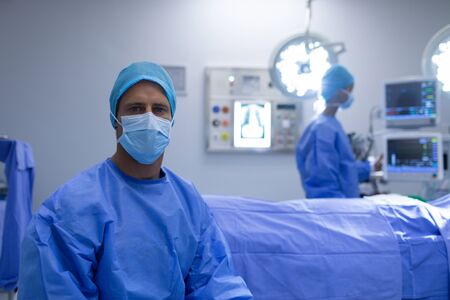 Front view of Caucasian male surgeon sitting with surgical mask in operation theater at hospital while African-american female surgeon working behind.の写真素材