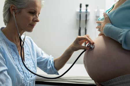 Side view of Caucasian female doctor examining pregnant Caucasian woman belly with stethoscope in examination room at hospitalの写真素材