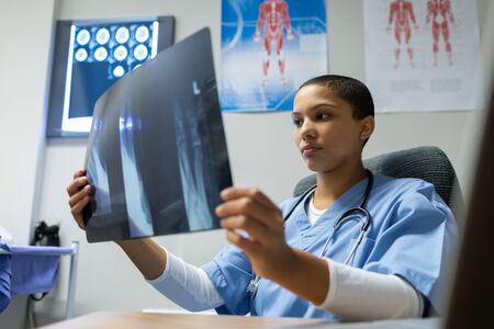 Front view of mixed-race female doctor examining x-ray report at desk of examination room in hospitalの写真素材