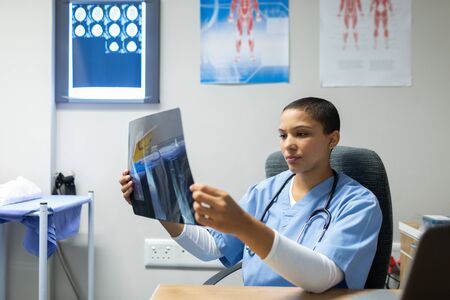 Front view of mixed-race female doctor examining x-ray report at desk of examination room in hospitalの写真素材