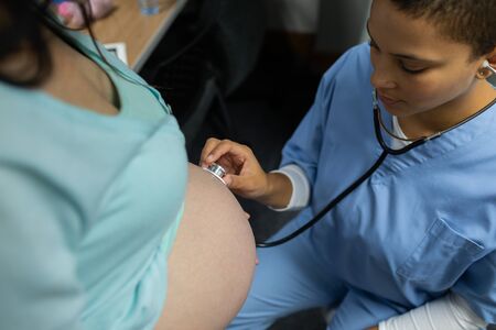 High angle view of Caucasian female doctor examining pregnant woman belly with stethoscope in examination room at hospitalの写真素材