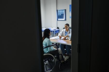 Front view of Mixed-race female doctor giving prescription to disabled female patient at desk in hospitalの写真素材