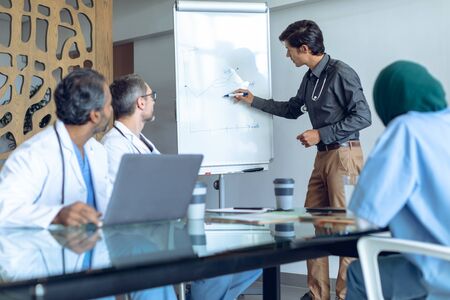 Front view of Caucasian male doctor explaining over flip chart in meeting at hospital. Coffee cup, medical folders, clipboard, digital tablet and laptop are on the table.の写真素材
