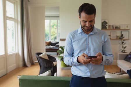 Front view of happy Caucasian man using mobile phone in a comfortable homeの写真素材
