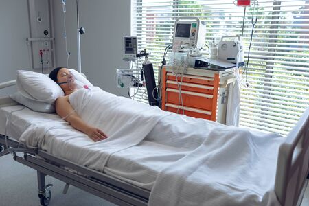 Front view of Asian female patient resting with oxygen mask and iv drip in bed in ward at hospital