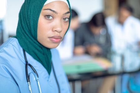 Side view of young mixed race female surgeon in hijab looking at camera in the hospital. With colleagues in the backgroundの写真素材