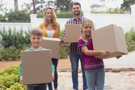 Front view of happy Caucasian family holding cardboard boxes in home yardの写真素材