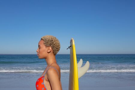 Side view of beautiful African american woman in swimwear standing with surfboard on beach in the sunshineの写真素材