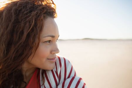 Side view of beautiful mixed race woman wrapped in blanket standing on beach in the sunshineの写真素材