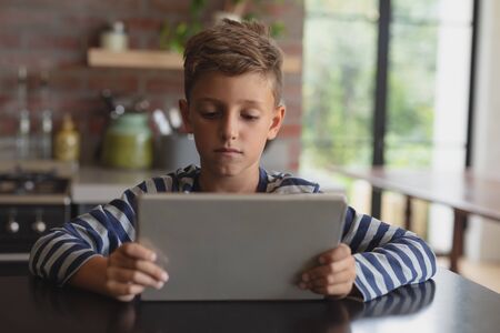 Front view of adorable Caucasian boy using digital tablet at table in kitchen at homeの写真素材