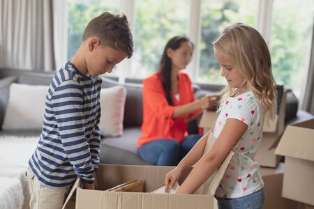 Side view of Caucasian siblings unpacking cardboard box while Caucasian mother looking a document in living room at homeの写真素材