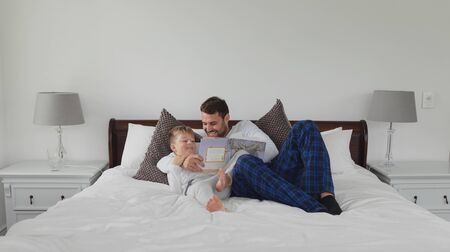 Front view of Caucasian father and son reading a story book while lying on bed in bedroom in a comfortable homeの写真素材
