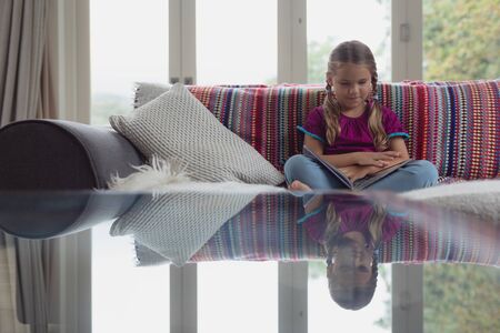 Front view of cute Caucasian girl reading a book on sofa in a comfortable homeの写真素材