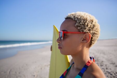 Side view of beautiful African american woman in bikini and sunglasses standing with surfboard on...