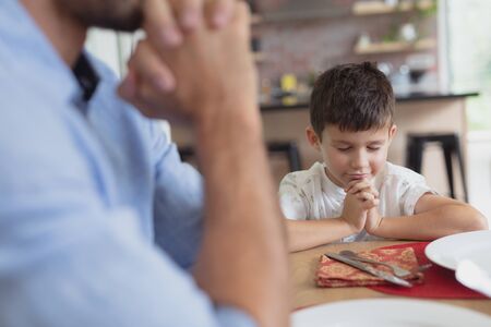 Side view of Caucasian father and son praying before having lunch at dining table in comfortable homeの写真素材