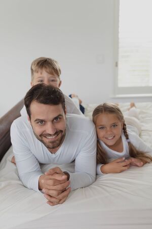 Front view of happy Caucasian father and children looking at camera while lying on bed in bedroom at comfortable homeの写真素材