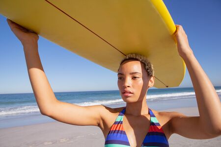 Front view of beautiful African american woman in bikini carrying the surfboard on her head at beach in the sunshineの写真素材