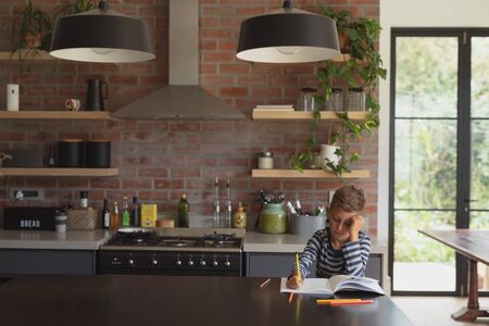 Front view of adorable Caucasian boy studying at table in kitchen at homeの写真素材
