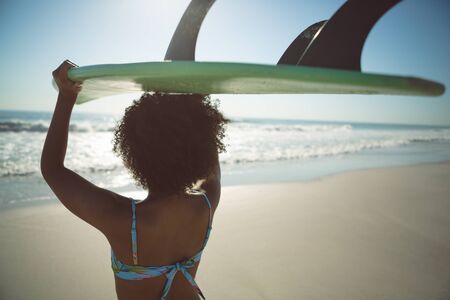 Rear view of African american woman carrying surfboard on her head at beachの写真素材