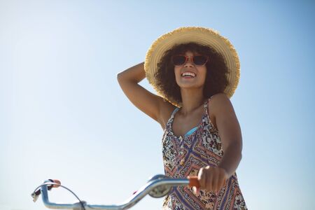 Low angle view of happy African american woman with bicycle at beach on a sunny dayの写真素材