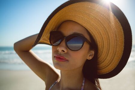 Front view of beautiful mixed race woman in hat and sunglasses standing on the beach