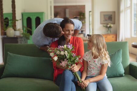 Front view of Caucasian daughter giving bouquet to mother while father kissing her in living room at homeの写真素材