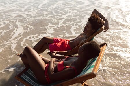 High angle view of African-american couple interacting with each other while relaxing in a beach chair on the beachの写真素材