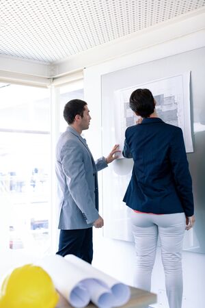 Rear view of Caucasian architects discussing over blueprint on glass board in a modern office