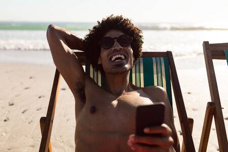 Front view of happy African-american man using mobile phone while relaxing in a beach chair on the beachの写真素材