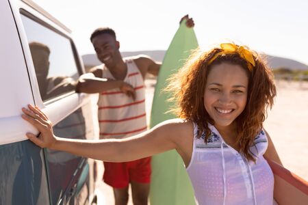 Front view of happy young Mixed-race couple with surfboard leaning on camper van at beachの写真素材