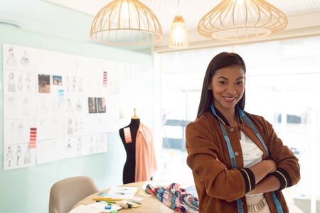 Front view of happy mixed race female fashion designer with arms crossed looking at camera in a modern officeの写真素材