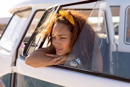 Side view of thoughtful young Mixed-race woman leaning on window of camper van at beach